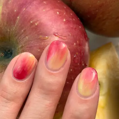 A hand with manicured nails near apples, nail art resembling apple skin coloration