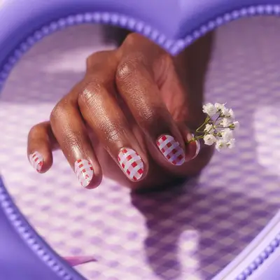 A hand with gingham patterned nails holding a small white flower, framed by a purple heart-shaped frame
