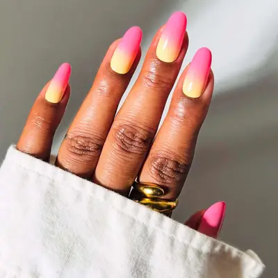 A hand with a gradient pink and yellow manicure, wearing gold rings