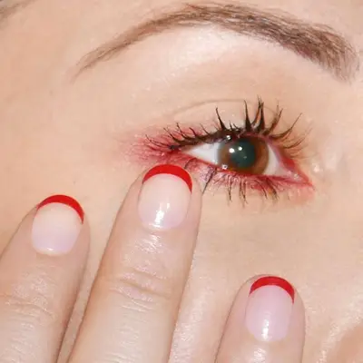 Model holding short nails with red French tips up against an eye makeup look with brown mascara and subtle red eyeshadow