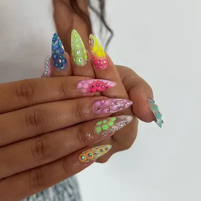 Hands showing long, elaborate fingernails decorated with gemstone designs and bright colors, intricate nail art patterns evident