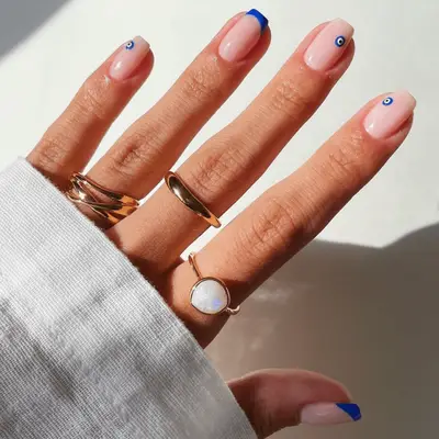 Short square-shaped nails with pink base and blue french tips and evil eyes on fair skin tone