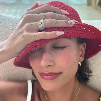 Hailey Bieber in a red crochet bucket hat displaying sheer pink chrome nails with strawberry decals.