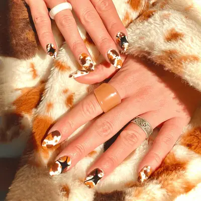Almond shaped nails with a brown, black, and orange cow print design and gold beads.