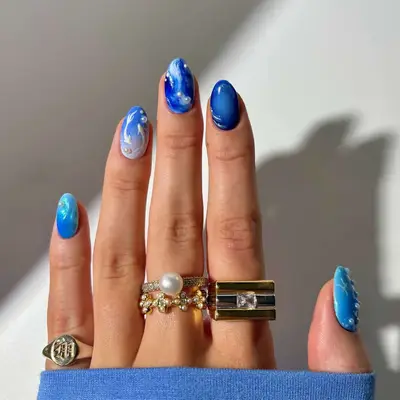 Hand with artistic blue and white nail designs and multiple gold rings, including one with a pearl
