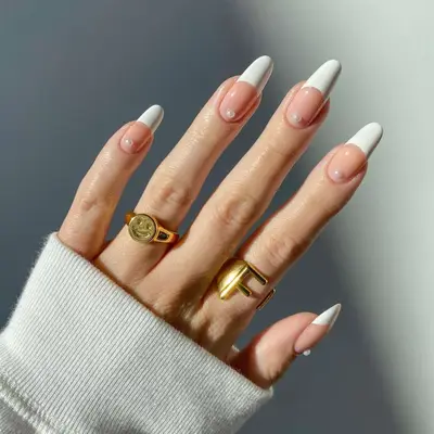 A hand wearing two gold rings displaying nails with a pearl-dotted French manicure design