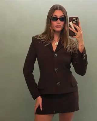 Hailey Bieber in a suit and sunglasses and strawberry chrome nails while taking a mirror selfie