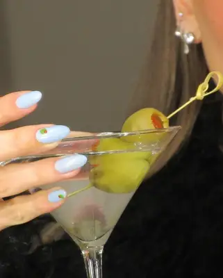 Hailey Bieber holding a martini with olives showing her blue manicure with olive design on a few nails