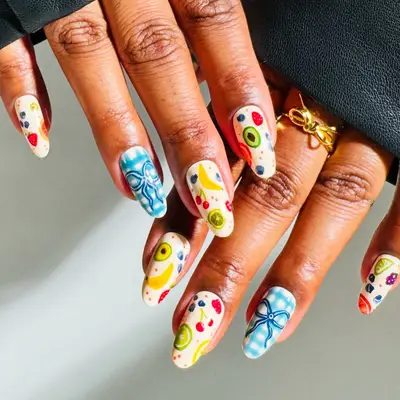 Hands with colorful nail art featuring fruit and bow designs, wearing a ring
