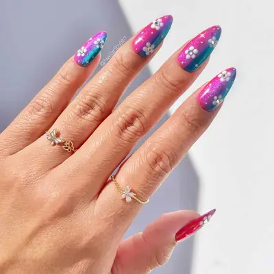 close up of nails with half of nail in pink, middle purple, other half blue, sparkles with white flowers and rhinestones