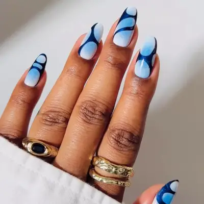 A hand with oval nails styled with a blue chrome gradient and abstract patterns, wearing gold rings and a white sleeve