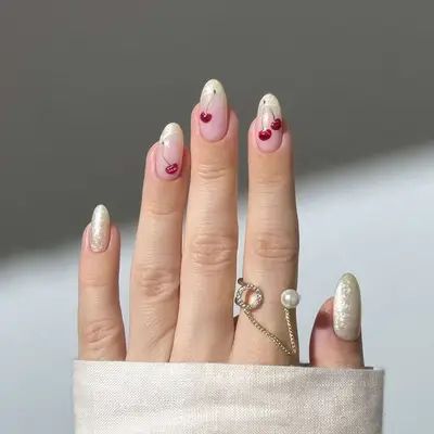 Hand with pearly nails featuring small red cherry designs, wearing thin gold and pearl rings