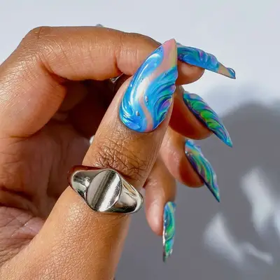A hand displayed with long, curved nails having blue and green wave designs, and a silver ring on the index finger