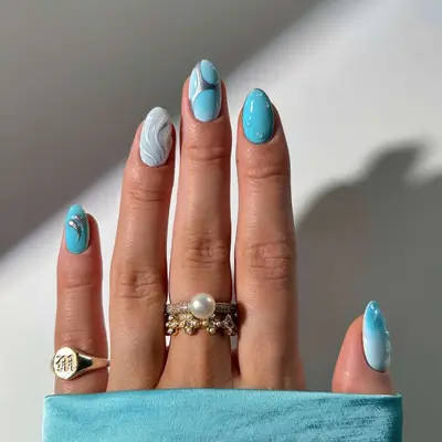 A hand with painted fingernails featuring a mix of patterns and shades of blue, jewelry on the fingers