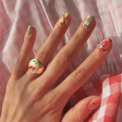 A hand with fruit-themed nail art and a ring with a cherry motif on a checkered fabric background