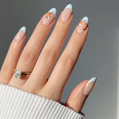 A hand displaying manicured nails with light blue tips and small orange designs on some nails, wearing a white top and a ring