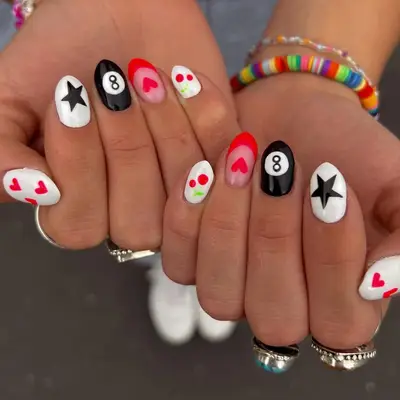 A hand with mixed nail art designs, including hearts, cherries, stars, and the number eight on pool balls
