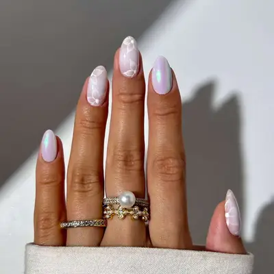 A hand with holographic white floral chrome nails, adorned with multiple gold and pearl rings
