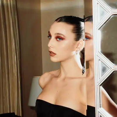 EMma chamberlain looking into the mirror with a black dress, slicked back hair and cherry red eye makeup