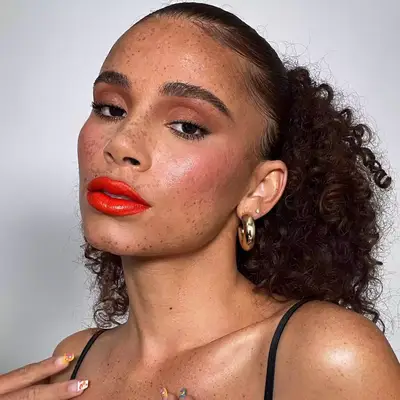 Joyjah Estrada wears a warm-toned dewy makeup look with bright red-orange jelly lips