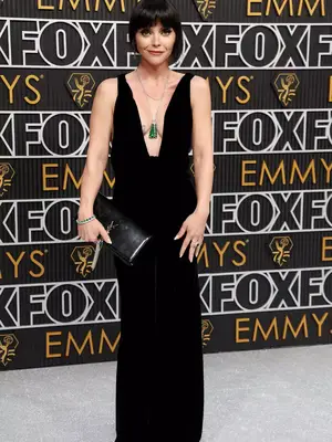 Christina Ricci wears a black gown with plunge neckline, YSL clutch, and layered silver necklaces with green pendant to the January 2024 Emmy Awards