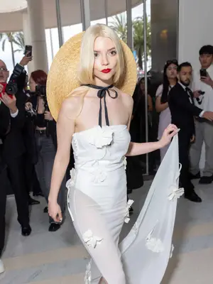 Anya Tayloy-Joy wearing a white sheer dress with a large hat tied around her neck and bright red lipstick.