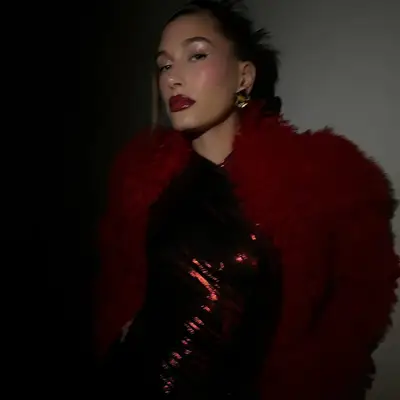 Hailey Bieber in frosted chrome eye makeup, cherry red lips, a red fur coat, and metallic red dress