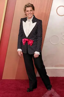 Brandi Carlile wears a black and white polka-dotted jacket with red bow tie closure, black pants, and white blouse to the 2025 Oscars