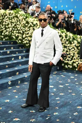 Pharrell Williams wears a white pinstriped jacket, black flared trousers, sunglasses, and white shirt with black tie to the 2025 Met Gala