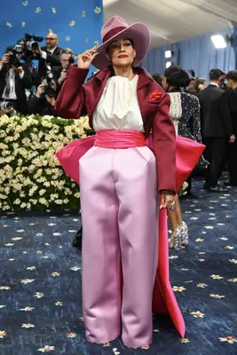 Tracee Ellis Ross wears a red and pink suit with asymmetrical hat, oversized bow cummerbund, wide leg pants, and puffy blouse to the 2025 Met Gala