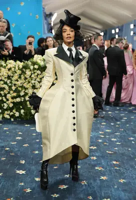 Tessa Thompson wears a structured cream coat, asymmetrical sparkly black top hat, and platform boots to the 2025 Met Gala
