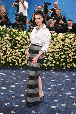 Anne Hathaway wears a structured white blouse, black and gray striped maxi skirt, elaborate jewelry, and high ponytail hairstyle to the 2025 Met Gala