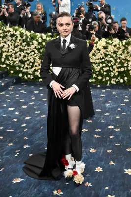 Aimee Lou Wood wears a cropped suit jacket with white corsage, dress with train, flower-covered Mary Janes, and silver jewelry to the 2025 Met Gala