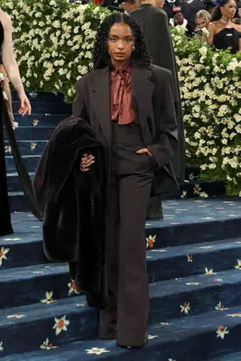 Yara Shahidi wears an oversized suit with rust-colored blouse and carries a velvet coat to the 2025 Met Gala