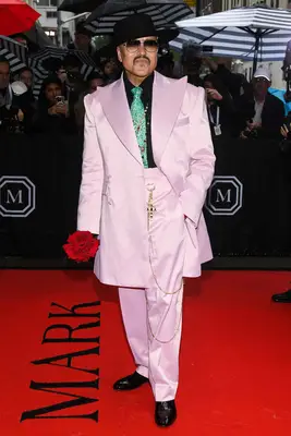 Willy Chavarria wears a pink suit with green tie, black hat, and red flower to the 2025 Met Gala