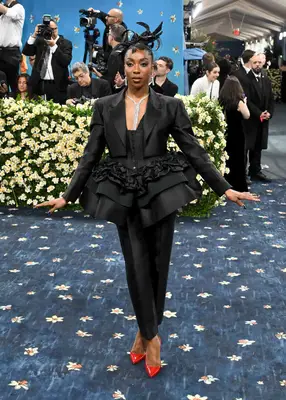 Ego Nwodim wears a peplum suit jacket, trousers, sculpted hairstyle, sparkly jewelry, and red pointed-toe shoes to the 2025 Met Gala