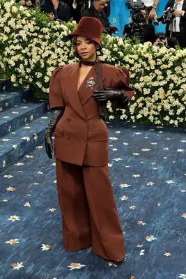 Kara Young wears a voluminous chocolate brown suit with black leather opera gloves, tie as scarf, and bucket hat at the 2025 Met Gala