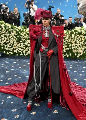Teyana Taylor wears a black and red pinstriped suit, red vest, silver chains, and red cape with florals and feathered hat to the 2025 Met Gala