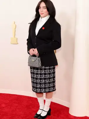 Billie Eilish wears a black blazer, white button-down, tweed skirt and purse, and Mary Janes with white socks to the 2024 Oscars