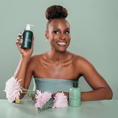Issa Rae sitting at a table holding two haircare products with pink flowers.