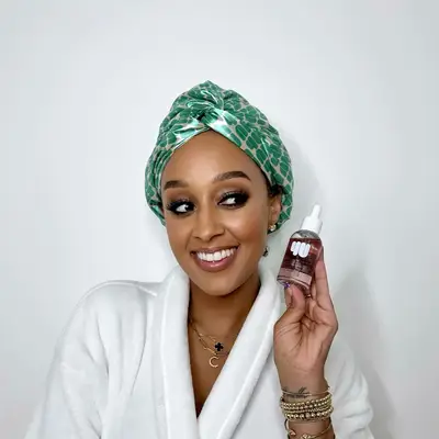 Tia Mowry smiling while holding a hair oil from her haircare brand, 4U by Tia.