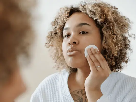 A woman in a robe applying toner to her face with a cotton pad
