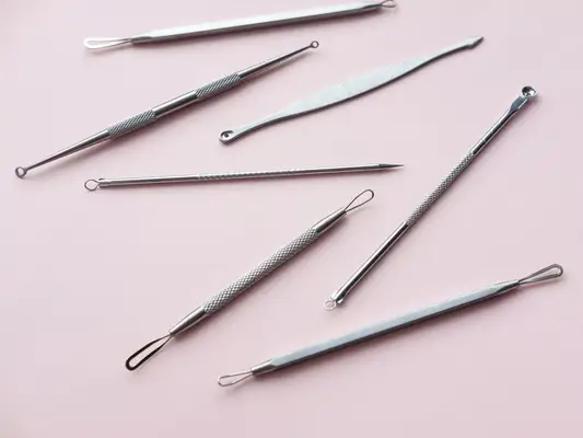 Close up of multiple steel acne extraction tools laid out on a light pink background.