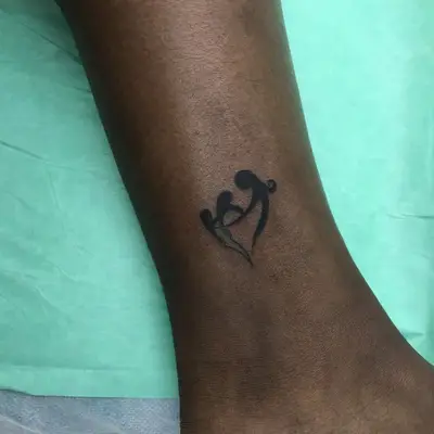 Small ankle tattoo of figures holding hands in a heart shape