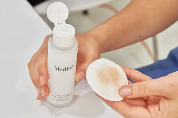 Hands holding a bottle of Medik8 Eyes & Lips Micellar Cleanser and a used cotton round