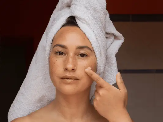 A woman with her hair in a towel and applying skincare to her face with one finger