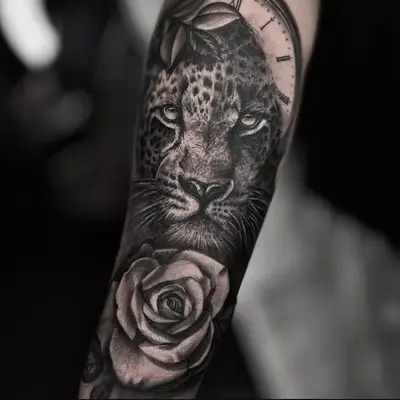 A grey and black tattoo of a leopard, clock face, and rose on the lower arm.