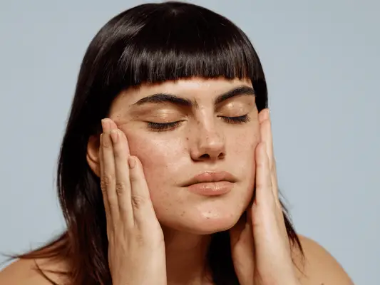 Model with medium length brown hair, blunt bangs, and freckles holding her face.