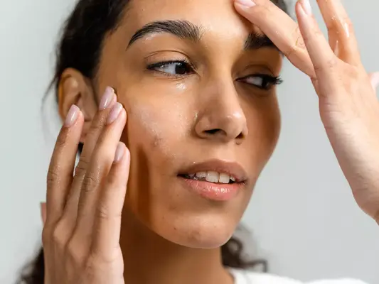 Close up of a model looking off camera, applying clear product to her face with both of her hands.