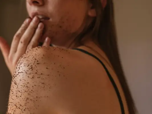 A woman with a skincare scrub on her shoulder, face, and hands, touching her fingers to her lips